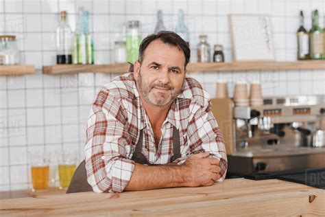 Happy Mature Bartender At Cafe Leaning On Bar Counter And Looking At