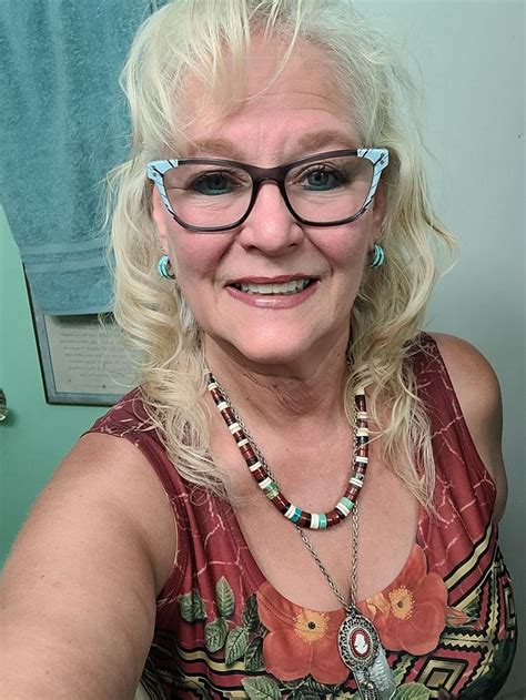 An Older Woman With Glasses Sitting In Front Of A Bathroom Mirror Smiling At The Camera