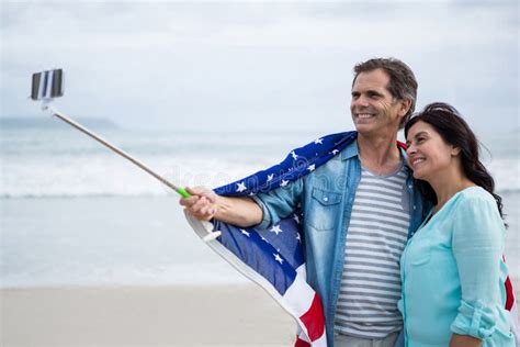 Couple Wrapped In American Flag Taking Selfie On Beach Stock Image