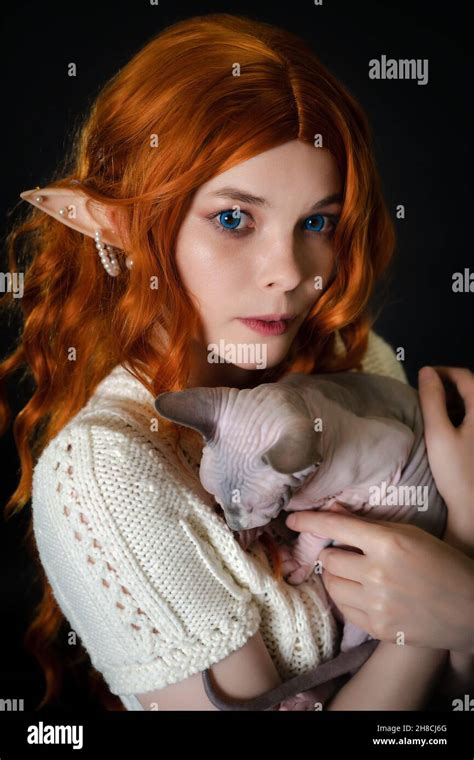 Portrait Of Cosplay Elf Young Woman In White Knitted Dress Holding Sleepy Sphinx Cat In Hands On