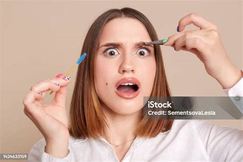 Portrait Of Shocked Scared Female In Beauty Salon Holding Eyebrow Brush And Tweezers Looking At