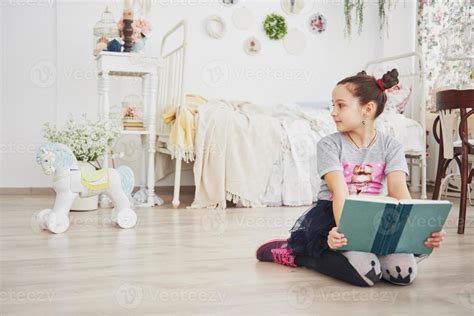 Cute little child girl reading a book in the bedroom. Kid with crown sitting on the bed near ...