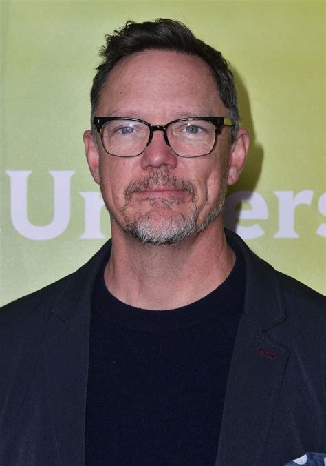 a man with glasses standing in front of a green wall wearing a black ...