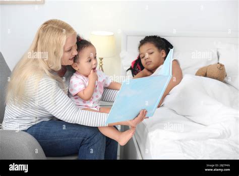 Female Mature Nanny Reading Book To Little African American Sisters In