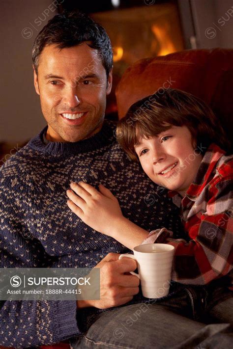 Father And Son Relaxing With Hot Drink Watching TV By Cosy Log Fire SuperStock