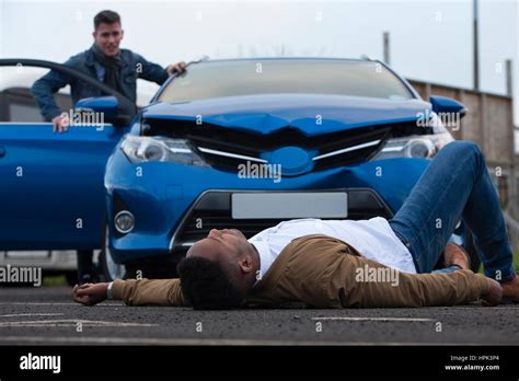 A Man Is Lying In Front Of A Damaged Car Which Has Just Hit Him The