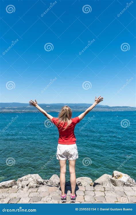 View From Behind Of A Blonde Unrecognizable Tourist With Her Arms Raised In Front Of Lake Geneva