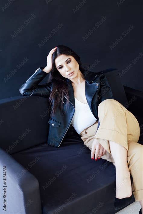 Beautiful Brunette Girl Sitting On The Couch In A Dark Room On A Black Background Stock Photo