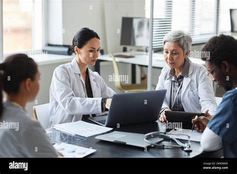 Brunette Female Clinician In Lab Coat Pointing At Laptop Screen While Making Presentation Of