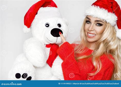Beautiful Blonde Female Model Dressed In A Santa Claus Hat Embraces A White Teddy Bear In A Red