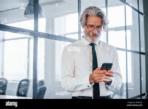 Mature Businessman With Grey Hair And Beard In Formal Clothes Is In The Office With Phone In