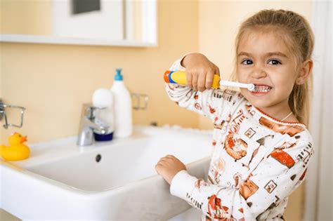 Une jolie petite fille blonde se brosse les dents avec une brosse électrique debout dans la