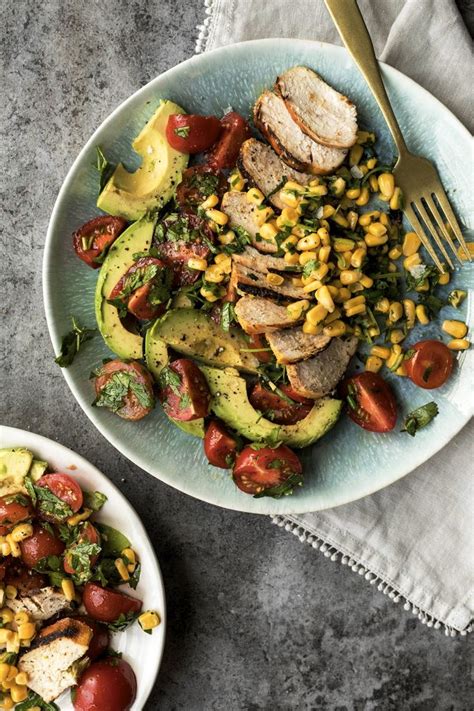 A White Plate Topped With Meat And Veggies Next To A Bowl Of Corn