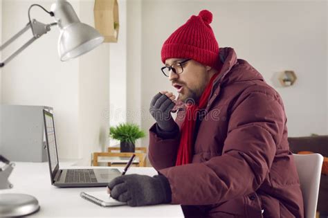 Freezing Man Sitting At Desk Blowing In His Hands Trying To Keep Warm