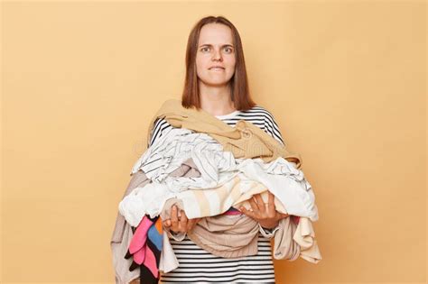 Confused Brunette Woman Holding Pile Of Clothes In Hands Isolated Over Beige Background Looking