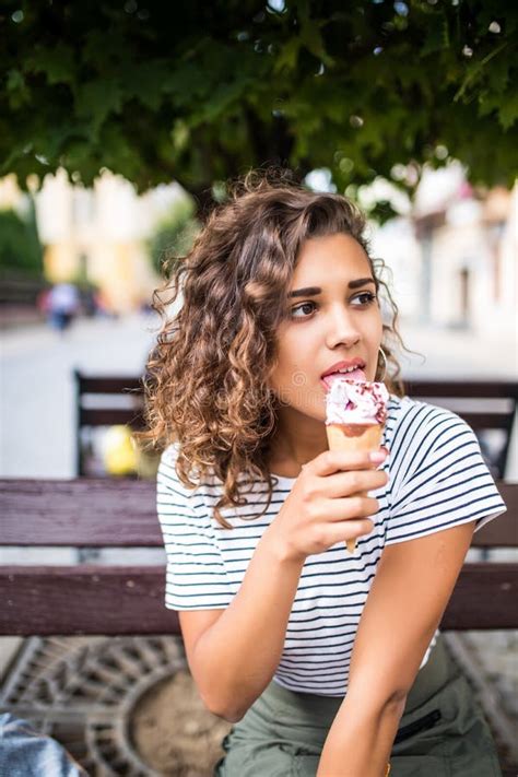 Mujer Rizada Latina Joven Que Se Sienta En Un Banco De Parque Y Que Come El Helado En El Verano