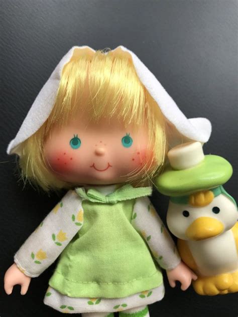 A Doll With Blonde Hair And Green Dress Holding A Toy Penguin In Front