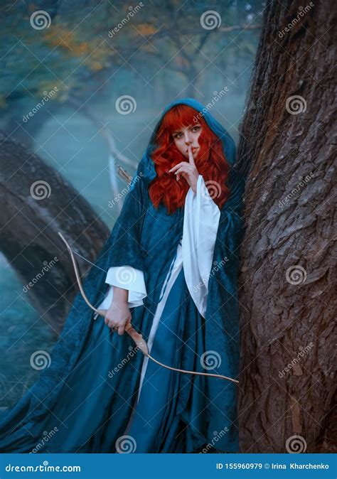 Red Haired Girl In Blue Cloak Standing By Tree And Looking Directly At