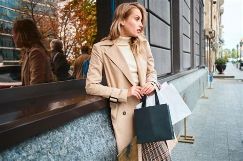 Vue latérale d une élégante fille blonde en manteau beige avec des sacs à provisions regardant