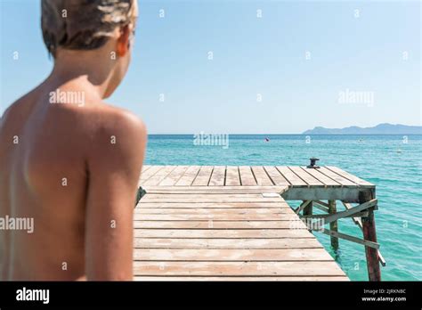 Back View Of Teenage Boy With Naked Torso And Wet Hair Standing On Wooden Quay Near Blue Sea On