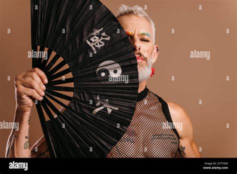Bearded Mature Gay Man With Colored Eyebrows Covering Half Face With Fan Against Brown