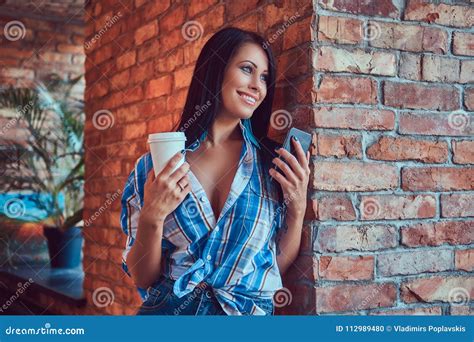 A Happy Brunette In A Flannel Shirt And Jeans Holds A Cup Of Coffee And The Phone Posing Against