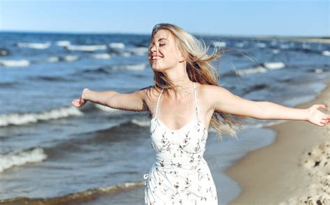 Happy Blonde Beautiful Woman On The Ocean Beach Standing In A White Summer Dress Raising Hands