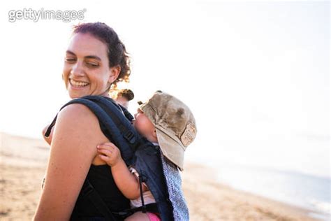 Smiling Mother Carrying A Baby With A Cap On Her Back In A Baby Carrier