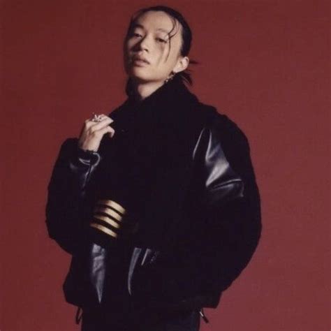 A Man Standing In Front Of A Red Wall Wearing A Black Jacket And Gold Cuffs