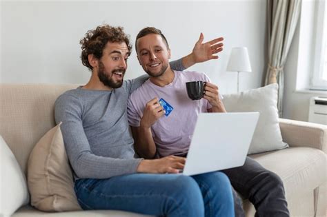 Alegre joven pareja gay sonriendo alegremente mientras compra en línea en casa dos jóvenes