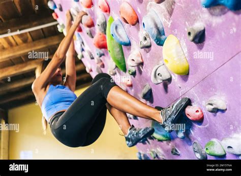 From below full body sportswoman in activewear hanging on grips of tilted climbing wall over ...