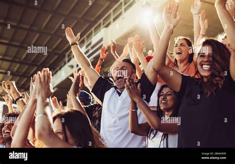 Crowd of sports fans cheering during a match in stadium. Excited people standing with their arms ...