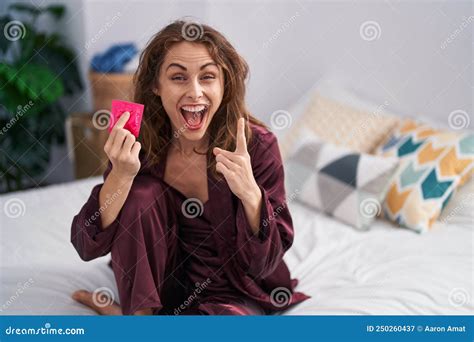 Beautiful Brunette Woman Holding Condom At The Bedroom Surprised With An Idea Or Question