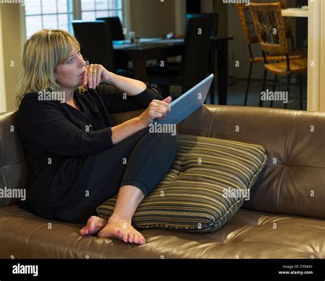 Femme blonde est assise sur son canapé en cuir avec une tablette et regarde au loin dans l