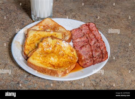 A Plate Of French Toast That Has Powdered Sugar And Maple Syrup On It With Bacon And A Glaso Of