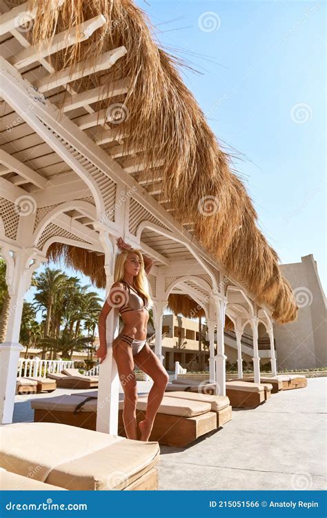 Woman Wearing Bikini Standing At Lounger Under Straw Canopy At Resort