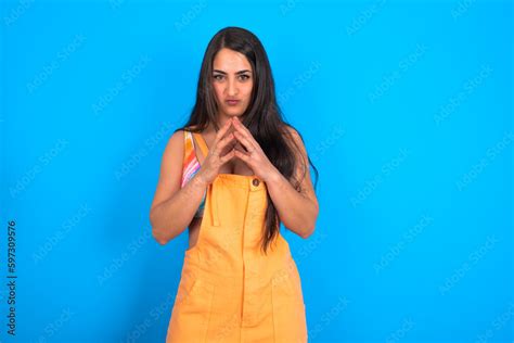 brunette woman wearing orange overalls over blue studio background ...