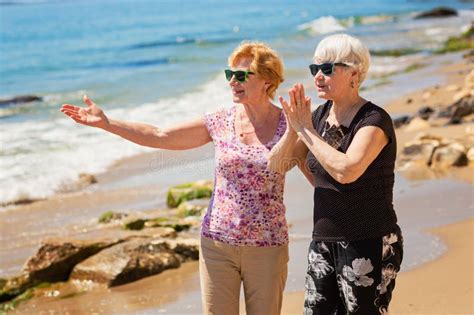 Two Senior Women Are Walking Along The Rocky Shore Talking And