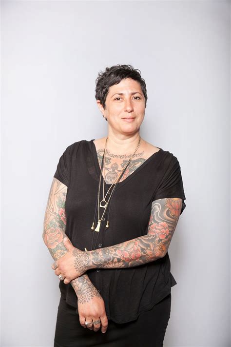 A Woman With Tattoos Standing In Front Of A Gray Wall Wearing A Black