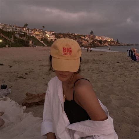 A Woman Sitting On Top Of A Sandy Beach Next To The Ocean Wearing A Hat