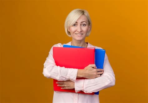 Lächelnde junge blonde slawische frau ordner lokalisiert auf orange wand hält Kostenlose Foto