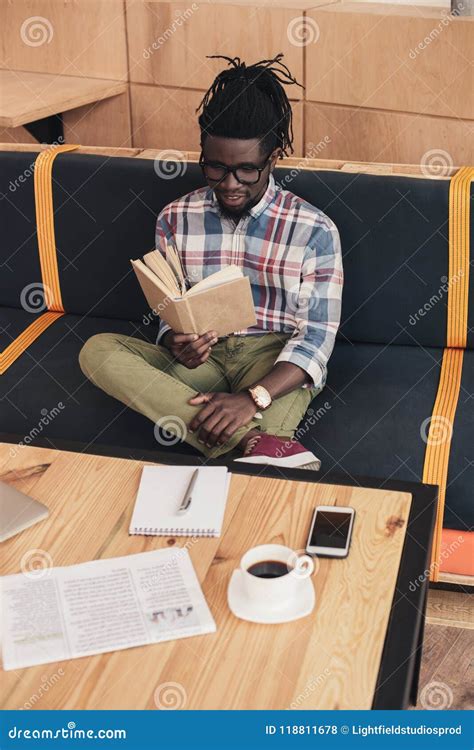 African American Man Reading Book While Sitting On Sofa Stock Photo