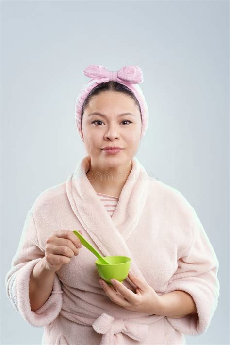 Mature Asian Woman Dressed In Bathrobe Holding Cosmetics In Her Hands While Standing Against