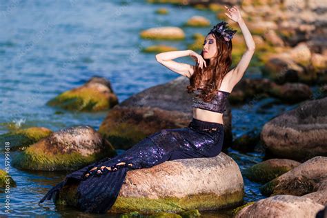 Asian Mermaid Posing Near Ocean Shore On Rocks While Wearing Seashell