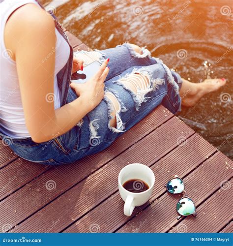 Young Woman Using Her Smartphone Sitting On The Pier And Drinking Hot