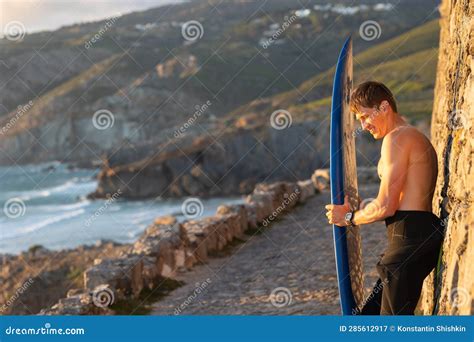Fit Smiling Man With Naked Torso Standing By The Wall Holding A Surfboard At Early Sunset Stock