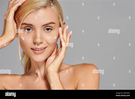 Portrait Charming Blonde Girl With Her Hands Around Head And Cream On Cheeks Against Grey