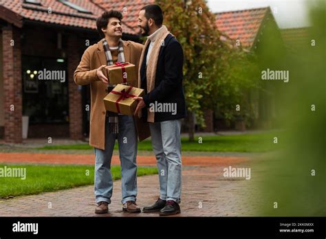 Full Length Of Happy Gay Couple In Trendy Outfits Standing With Gift Boxes On Blurred Foreground