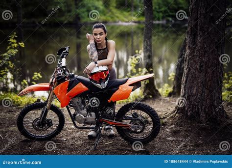 Charming Tattooed Racer Girl Wearing Motocross Outfit With Semi Naked Torso Leaning On Her Bike