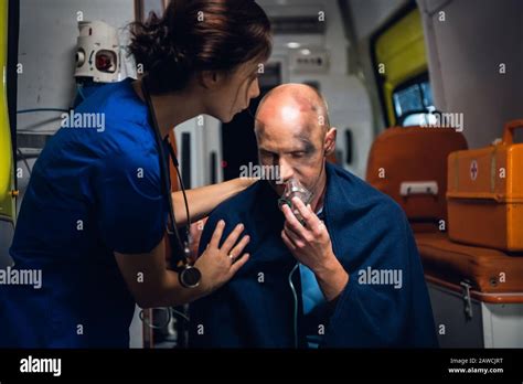 Woman In Blue Medical Uniform Stands With Man Who Sits In Oxygen Mask